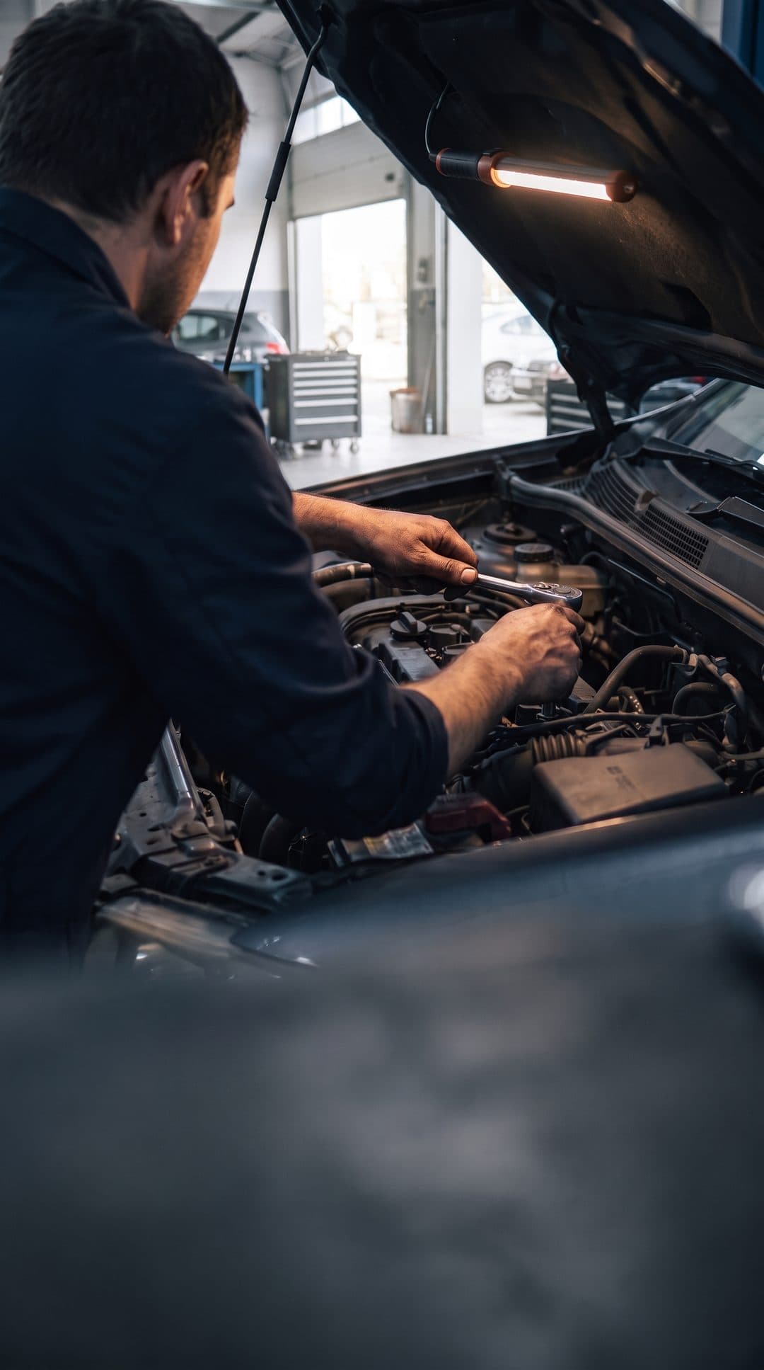 Mechanic seen from behind, hands in the engine of a car with the bonnet up, in a workshop